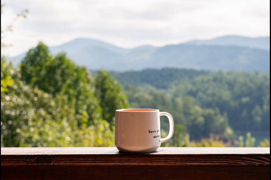 White mug resting on a wooden ledge with forest and mountains beyond, suggesting a quiet pause and reflection.